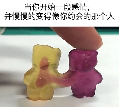 图片上方配有文字'当你开始一段感情，并慢慢的变得像你约会的那个人'，下方为实物拍摄图：两只小熊软糖（黄色和紫色）的手臂部分相互融合在一起，一只手指正按压在紫色小熊软糖的头顶。整体通过小熊软糖的融合状态，比喻恋爱关系中双方逐渐变得相似的现象，画面生动且富有生活气息。
