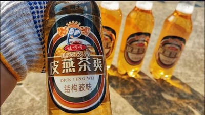 画面中一只戴着带蓝色防滑点劳保手套的手，拿着一瓶模仿娃哈哈包装风格的恶搞饮料，背景还摆放着三瓶同款饮料。饮料包装上标注“娃哈哈 迪克饮品 皮燕茶爽”，口味是离谱的“结构胶味”，其中“皮燕”是“屁眼”的谐音，属于玩谐音梗的整活恶搞内容，整体风格搞笑又离谱，是网友制作的不存在的恶搞饮品图。