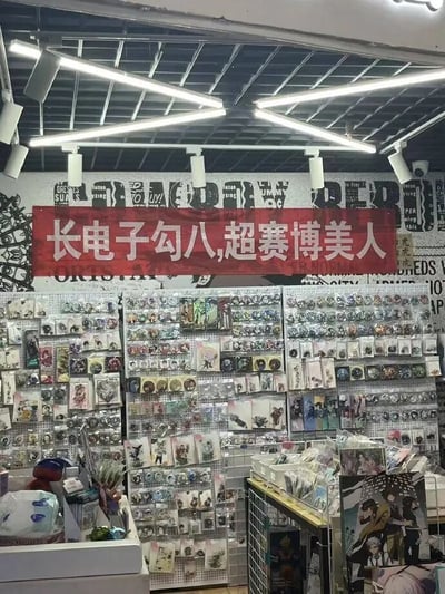 一家动漫周边商店的内部场景，货架上摆满了各种动漫徽章、卡片和周边产品。店内灯光明亮，采用工业风装修，天花板有裸露的金属结构和长条形灯管。最引人注目的是墙上悬挂的红色横幅，上面写着“长电子勾八,超赛博美人”的标语，其中“勾八”是较为粗俗的方言词汇，与“超赛博美人”形成幽默对比。