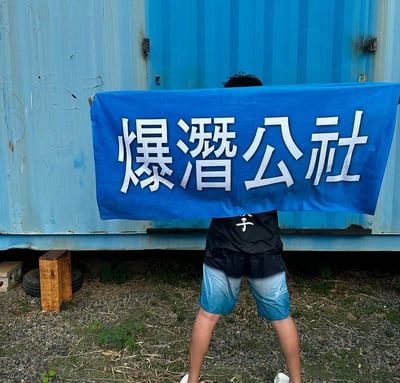 图片中一个人站在蓝色金属集装箱前，用一块蓝色横幅挡住了自己的脸。横幅上用白色大字写着'爆潛公社'。此人穿着黑色上衣和蓝白渐变短裤，站在铺着碎石的地面上，旁边有一些杂草和一个木箱。