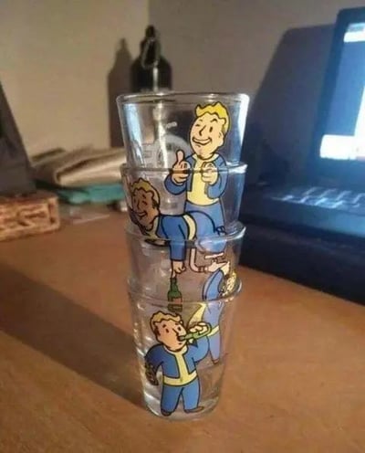 一张展示《辐射》游戏经典角色Vault Boy主题酒杯的照片。四只透明玻璃杯叠放在木质桌面上，每只杯子上都印有不同姿势的Vault Boy：从上到下分别是竖大拇指点赞、侧身躺卧、手持酒瓶和举杯饮酒的造型。背景可见打开的笔记本电脑、堆叠的纸张和编织篮，营造出居家或办公桌上的休闲氛围。