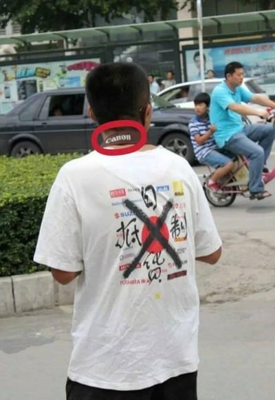 这是一张街头实拍照片，画面中的男生背对着镜头，穿着一件印有“抵制日货”字样的白色T恤，T恤上还印有多个日本品牌标识并被划掉，包括松下、索尼、铃木、本田、东芝等。但极具反差感的是，他脖子上的颈枕清晰印有日本相机品牌“canon（佳能）”的英文标识，形成了口号与实际行为完全矛盾的尴尬场景，充满了讽刺意味，用来调侃那些言行不一的人。