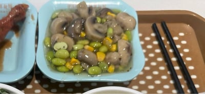 这是一张日常用餐场景的实拍图，画面核心是盛放在浅蓝色方盘中的清炒时蔬，食材包含对半切开的口蘑、鲜绿色的毛豆粒、明黄色的甜玉米粒，食材浸在清淡的调味汤汁中，色泽清新。左侧浅蓝色小盘中残留着带酱汁的肉类，右下角的棕色波点托盘上摆放着一双黑色筷子，画面底部还能看到其他餐品的边缘，整体呈现出家常、清淡的饮食氛围，给人清爽可口的感觉。