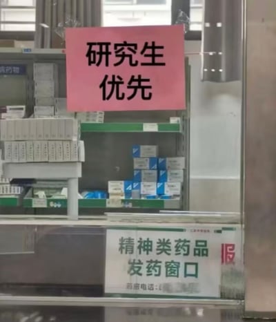 这是一张医院药房窗口的实拍图，玻璃上方贴着一张醒目的粉色标语，写着‘研究生优先’；玻璃下方的标识牌明确标注这是‘精神类药品发药窗口’，背景的货架上摆放着各类盒装药品。
这张图的反差感十足，将‘研究生’群体与‘精神类药品窗口’绑定，用黑色幽默的方式调侃了研究生群体普遍面临的精神压力大的现状。