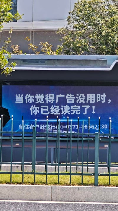 图片展示了一块户外广告牌，背景为蓝色，上方用白色醒目字体写着“当你觉得广告没用时，你已经读完了！”，下方附有文字“留住客户赶快行动：0371-88562130”。广告牌被绿色金属围栏遮挡部分，周围有树木和现代建筑，呈现出城市户外场景。
该广告运用了巧妙的心理暗示：当观众产生“广告没用”的想法时，实际上已经完整阅读了广告内容，以此证明广告的有效性，形成有趣的反转效果。