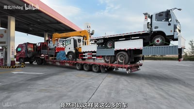 图片展示了加油站场景，一辆红色底盘的大型平板运输卡车停在加油站内，车身上分层装载了多辆工程车辆，包括一辆黄色起重机卡车和一辆白色货车，车辆通过绳索固定。加油站顶棚为红白色，背景是蓝天白云。图片底部配有文字“竟然可以这样装这么多的车”，直观呈现了高效的车辆装载方式，给人以运输能力强大的视觉冲击。