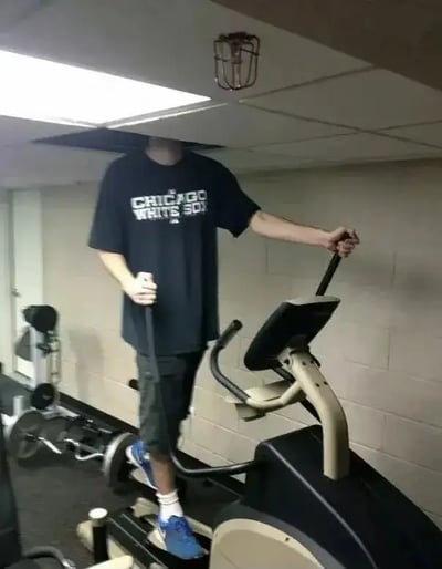 这张图拍摄的是一个穿着“CHICAGO WHITE SOX”T恤的高个子男子在健身房踩椭圆机，但因为房间高度太低，他的头直接顶进了天花板，形成了头部消失的有趣视觉效果。画面自然幽默，带有物理错位和生活尴尬双关，适合表达“太高了不适用”或“小空间心酸”等情境。