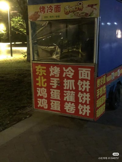 这是一张夜晚拍摄的街头移动小吃餐车实拍图，餐车停在铺有地砖的路边，背景可见路灯灯光和绿植。餐车顶部为黄色招牌，标明售卖烤冷面和手抓饼，下方红色背景的菜单采用四行四列的排版，若按竖列读取第一列的文字会产生“东北鸡鸡”的搞笑歧义，实际正确读取方式为横向逐行阅读，分别对应东北烤冷面、东北手抓饼、鸡蛋灌饼、鸡蛋卷饼四款餐品，图片右下角带有小红书平台的水印和发布者的账号ID，因菜单排版造成的意外笑点在网络上传播。