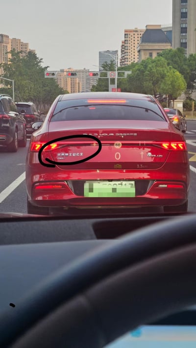这是一张从车内视角拍摄的城市道路车流照片，画面主体是一辆红色比亚迪秦PLUS轿车的车尾，车主将原本的比亚迪标识创意改装为“比亚迪士尼公主”，玩了“比亚迪”与“迪士尼”的谐音梗，同时保留了“DM-i”的标识。车尾上方的“BUILD YOUR DREAMS”和右侧的“秦PLUS”字样清晰可见，背景中能看到城市高楼、绿树以及亮起红灯的交通信号灯，显示当前车辆处于红灯等待通行的状态，周围还有其他待行车辆。
