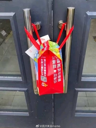图片展示了一个挂在双开玻璃门金属把手上的红色蜜雪冰城包装袋。袋子通过红色提手缠绕在两个门把手上，袋身印有白色文字“雪冰做甜蜜 我爱你 你爱我”及“雪王带你甜蜜蜜 乡村儿甜”，并配有一个二维码图案。背景可见门玻璃后的瓷砖地面，整体场景为日常街头的随拍画面，呈现出温馨可爱的氛围。