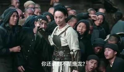 这是一张电影《龙门飞甲》的剧照截图，画面中周迅饰演的顾少棠手持武器，表情严肃地站在一群衣衫褴褛、面露惊讶的百姓中间。图下方被添加了玩梗字幕“你还说你不是唐龙？”，该字幕出自电影《功夫》的经典台词，将毫无关联的影视场景与台词结合，通过错位反差制造搞笑效果。