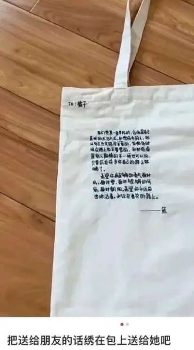 这是一张静态单图，属于原始拍摄图与图文混排的内容，展示了一个放置在浅棕色木质地板上的白色帆布袋，袋身用黑色绣线绣有一段送给名为“橘子”的人的暖心赠言。赠言开篇点明对方是独一无二的，无需刻意模仿他人，鼓励对方坚持走自己的路，不必焦虑；接着希望对方在面对人生中的风雨、低谷与美好时，都能自在地做自己，走属于自己的人生道路，最后带有署名。图片底部配有文字“把送给朋友的话绣在包上送给她吧”，为观众提供了一种充满心意的手工礼物创意，整体传递出真挚温暖的友情氛围。