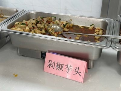 食堂 buffet 餐台特写，金属托盘内盛放着豆腐块、辣椒与酱汁混合的菜肴，托盘前立着粉色标牌，上书「剁椒芋头」四个大字。然而盘中食材明显为豆腐而非芋头，形成标签与实物不符的搞笑乌龙场景，可能是食堂工作人员误写菜名所致。