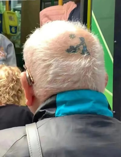 图片拍摄于公交车内部，聚焦一位白发 elderly man 的后脑勺。其头顶中央的 bald spot 上有一处黑色纹身，图案内容为一个小人正在推 Lawnmower，旁边还有一颗星星。背景中可见其他乘客的背影和公交车内的黄色扶手、绿色车门等设施，整体场景真实自然，因 elderly man 独特的头部纹身而显得格外有趣。