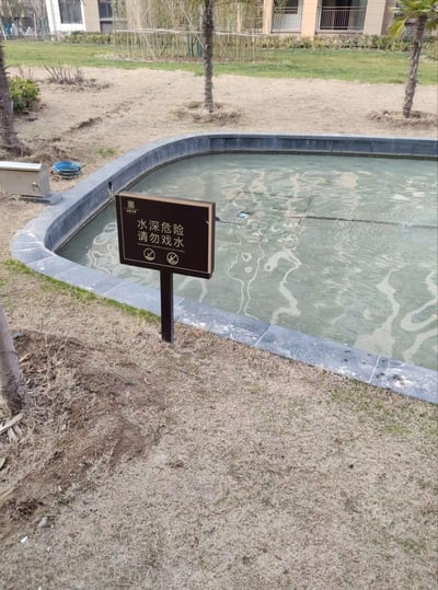 这是一张拍摄于小区内的实景照片，画面主体是一处水位极浅的景观水池，水面浑浊，能看到池底的情况。池边醒目地立着一块带有“品樾华府”标识的警示牌，上面写着“水深危险 请勿戏水”，还有对应的英文提示和禁止靠近、禁止戏水的图标。水池周边是枯黄的草地和几棵刚栽种的小树，背景可见小区住宅楼。实际浅水位和警示牌的“水深危险”内容形成强烈反差，显得十分滑稽，很可能是业主用来吐槽物业不合理的安全警示设置。