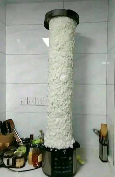 这是一张家庭厨房场景的实拍图，画面中心的家用电饭煲在完成煮饭操作后，内部的米饭因为米或水的投放量过多，加上加热过程中的膨胀效应，意外从电饭煲的上盖处大量挤出，形成了一根高耸的白色米柱，几乎顶到了厨房的墙面上方，造型十分夸张荒诞。周围的厨房台面上摆放着调料瓶、厨具收纳盒等日常厨房用品，背景是白色瓷砖墙和电源插座，普通的日常环境和夸张的米柱形成强烈反差，充满了搞笑的生活趣味，是典型的生活操作失误引发的搞笑场景。
