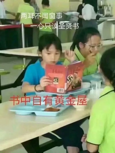 这是一张拍摄于学校食堂的实拍照片，后被添加文字制作成梗图。画面中心是一位穿着蓝色短袖的孩童，坐在餐桌前，面前摆放着装有少量食物的餐盘，正低头专注地捧着一本封面带有争议标志的书籍阅读；周围还有几位穿着绿色上衣的孩子在就餐。图片上方配文“两耳不闻窗事 一心只读圣贤书”，下方配文“书中自有黄金屋”，用传统的劝学诗句和孩童阅读的书籍形成强烈反差，制造出戏谑搞笑的效果，用来调侃这种专注读书的场景与书籍内容的不协调感。
