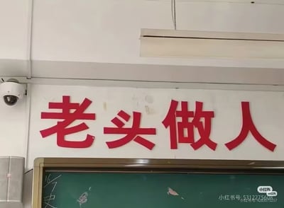 教室场景照片，白色墙壁上悬挂着红色立体字标语“老头做人”，标语下方是绿色黑板，黑板上有少量模糊粉笔痕迹。左侧墙壁安装有白色监控摄像头，标语上方为白色空调挂机。画面右下角有小红书平台用户ID水印。标语“老头做人”疑似因字形相近或发音戏谑产生的幽默改编，实际可能想表达“老实做人”等正面含义，形成反差笑点。