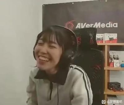 这是一张直播画面的截图，画面中的女主播戴着黑色头戴式耳机，身穿灰黑拼接的运动款上衣，正仰头开怀大笑，表情十分爽朗。背景可见带有「AVerMedia」（圆刚科技，专注于直播、影音设备的品牌）标识的黑色背景板，还有电竞椅与摆放着物品的置物架，整体是典型的个人直播空间场景，画面氛围轻松愉快。