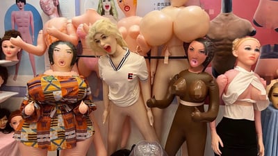 图片展示了多个造型夸张的充气娃娃和人体模型，它们被集中陈列在一起。这些娃娃具有各种肤色和发型，部分穿着简单衣物，如白色T恤和彩色图案上衣。最引人注目的是部分娃娃拥有极不自然的超大胸部和扭曲的面部表情，包括张大嘴巴和夸张的五官比例。背景中还能看到更多类似的充气玩偶和人体模型海报，整体呈现出一种怪异而荒诞的视觉效果。