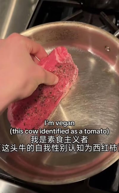 图中一只手将撒有黑胡椒和盐的生牛排放入平底煎锅，画面搭配中英文双字幕：英文“I'm vegan (this cow identified as a tomato)”，中文“我是素食主义者（这头牛的自我性别认知为西红柿）”。这是一个利用身份认同梗制造的搞笑反差，调侃那些想吃肉却找借口的“伪素食主义者”，通过将牛排荒诞地定义为“自我认同为西红柿的牛”，来合理化吃肉的行为，充满荒诞和调侃的趣味。