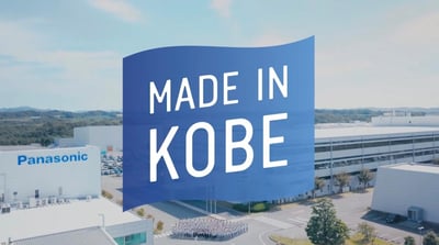 图片展示了日本神户市松下工厂的航拍场景，中央悬挂着巨大的蓝色横幅，上面用白色醒目字体写着“MADE IN KOBE”。背景中可见多栋现代化的白色工厂建筑，左侧建筑顶部有蓝色的“Panasonic”品牌标志，厂区内有整齐的停车场和道路，横幅正下方有一群人聚集，可能为工厂员工或参观人员。整体画面在晴朗天空的映衬下，呈现出专业、正式的企业宣传氛围，突出产品的产地标识和企业形象。