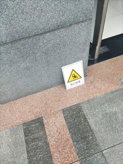 这是一张现实场景的实拍照片，画面中在建筑的灰色石材墙角旁，地面上立着一个带有白色边框的警示牌。警示牌主体是黄底黑图案的三角形警示标志，图案呈现出坠落的人形，下方配有中文文字“当心坠落”，用于提示此处存在坠落风险。地面是红灰拼接的石材地砖，旁边可见一处建筑出入口，整体场景是在室外公共场所，用于给路过的行人或进出建筑的人做安全提醒，图中没有双关、谐音梗或暗示内容。