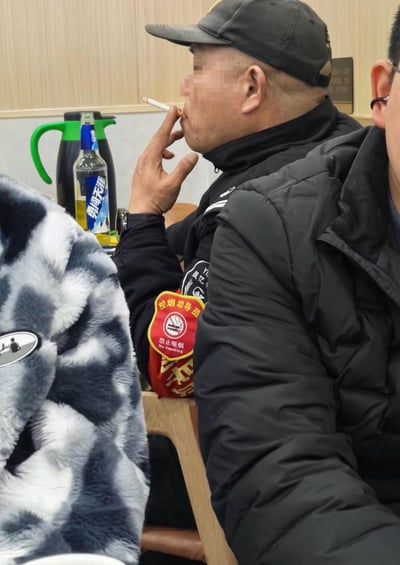 这是一张实拍的室内场景图，画面中心的中年男子头戴黑色鸭舌帽、身穿黑色外套，左臂佩戴着印有禁止吸烟标识、写有“控烟劝导员 禁止吸烟 No Smoking”字样的红色工作袖章，却正坐在椅子上用手指夹着香烟凑近嘴边吸烟，男子面前的桌上摆放着一瓶勇闯天涯啤酒和绿把手的黑色保温壶，画面左侧可见穿着蓝白毛绒外套的人的部分身体，右侧是另一名穿黑色棉服的男子的部分身体，整个场景呈现出极大的反差讽刺感。