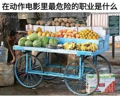 图片中展示了一辆装满西瓜、香蕉、菠萝、柑橘等多种水果的蓝色水果推车，一位摊主正弯腰在推车旁整理物品。图片上方配有文字“在动作电影里最危险的职业是什么”，通过幽默手法暗示：在动作电影的追逐、打斗场景中，水果摊主的推车常被撞翻破坏，因此水果摊主成了“最危险职业”的搞笑梗。