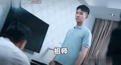 图片展示了《龙王赘婿》广告中的经典场景：一名身着浅蓝色polo衫的男子直立站在室内，背景可见电视和窗帘，前方有一人正低头向他鞠躬。画面中央配有文字“祖师”，精准捕捉了“卑微赘婿-身份揭露”的爽文套路关键时刻，暗示该男子隐藏的尊贵身份即将揭晓，为后续逆袭打脸情节铺垫。