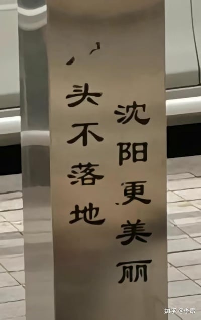 图片展示了沈阳街头一处立式标语牌，上面用黑色字体写着两行文字："沈阳更美丽 头不落地"。前半句"沈阳更美丽"是常见的城市宣传语，后半句"头不落地"则显得十分突兀且引人遐想。从字面看可能是提醒行人注意碰头的安全警示，但与前半句组合后产生了奇妙的幽默效果，引发网友趣味解读。背景可见街道地面和停放的车辆，显示这是在真实的城市环境中拍摄的。