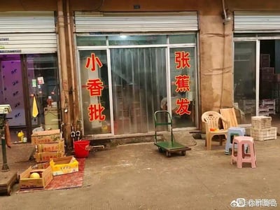 这是一张街边水果店的实拍图，店铺的玻璃推拉门两侧贴着红色竖排大字，左侧为“小香批”，右侧为“张蕉发”，实际是将“小香蕉批发”的字样拆分开来排版，利用汉字拆分制造出谐音梗效果，让原本普通的店铺招牌看起来像是两个奇怪的名称，充满搞笑趣味。店面前的水泥地面上，左侧堆放着装有水果的纸箱、红色水桶，还有老式秤具；中间位置有一辆绿色手推车；右侧摆放着塑料椅和几个不同颜色的小凳子，旁边还有白色塑料筐，整体呈现出普通街边小店充满生活气息的日常场景，而招牌的趣味设计成为画面的核心亮点。