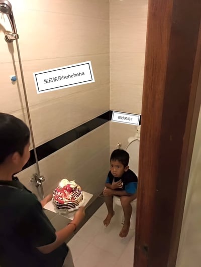 图片中，一名小孩坐在厕所马桶上，神情尴尬或不悦。另一人手捧生日蛋糕走进来，墙上配有幽默字体的文字：“生日快乐heheheha”，旁边配有小孩的回应文字：“很好笑吗？”。整个场景颇为滑稽，表现了生日庆祝与厕所尴尬场景的荒诞结合，带有一种无奈和自嘲的幽默感。该图片属于生活恶搞，适合用来调侃“我的生日也太离谱了吧”、“人生尴尬时刻”等网络语境。没有明显的谐音梗，但“heheheha”有调侃、嘲笑的情景意味。