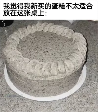 这是一张顶配底文的图文混排搞笑图，顶部配有文字说明，下方是实拍的灰色芝麻口味奶油蛋糕，蛋糕基底为灰色夹杂细小黑色芝麻颗粒，边缘有同色系奶油裱花，被放置在同样是灰色带黑色斑点的花岗岩桌面上，蛋糕的颜色、纹理和桌面高度相似，视觉上几乎要和桌面融为一体，形成了充满巧合的搞笑效果。