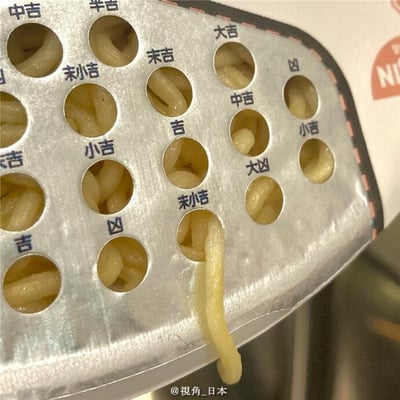 图片展示了一个金属托盘，托盘上分布着多个圆形孔洞，每个孔洞中都装有类似泡面面条的食物。孔洞周围用蓝色字体标注了类似神社签文的运势术语，包括“大吉”“吉”“中吉”“小吉”“末吉”“凶”“大凶”等。整体呈现出将传统 fortune-telling 签文与食物创意结合的趣味场景，给人一种新奇幽默的感觉。