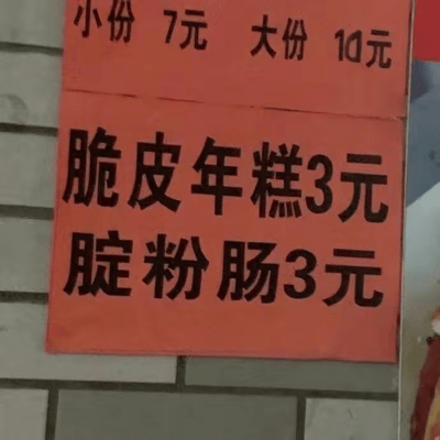 图片展示了一个街头小吃摊的手写价格牌，由两张橙色纸张组成，张贴在灰色砖墙背景上。上方纸张写着“小份 7元 大份 10元”，下方纸张则列出两种小吃：“脆皮年糕3元”和“腚粉肠3元”。

其中“腚粉肠”可能是摊主的笔误，“腚”字在中文里意为臀部，推测原本想写“淀粉肠”（常见的街头小吃）或“肠粉”，这种错别字意外形成了幽默效果，让普通的价格牌产生了趣味性。
