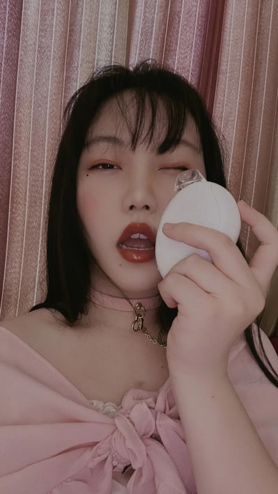 一位年轻女性正在进行化妆，她手持白色美妆蛋靠近脸部，单眼微闭，神情专注。她穿着粉色露肩上衣，佩戴着带有金属链条装饰的粉色项圈，妆容精致，涂着鲜艳的红色口红。背景是粉色条纹窗帘，整体色调柔和，营造出温馨的美妆氛围。