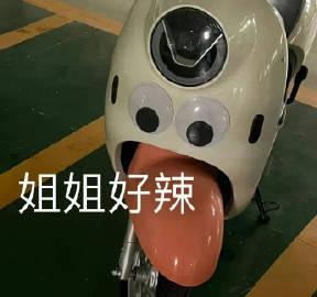 图片展示了一辆停在绿色地面（带有黄色停车线）的白色 scooter。 scooter 前部被创意改造成拟人化的脸：大型圆形大灯作为额头，两个贴有黑色圆心的圆形贴纸模拟眼睛，橙色的前挡泥板则形似伸出的舌头，表情俏皮。图片中央叠加了白色文字“姐姐好辣”，通过拟人化手法将 scooter 形容为“辣”（既指造型亮眼，也带有网络流行的夸赞意味），整体风格幽默可爱。
