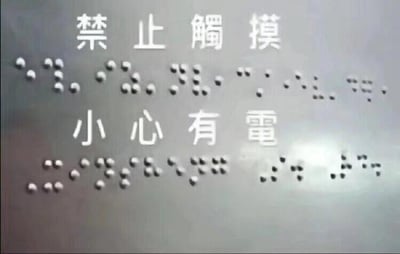 图片展示了一块警示标语牌，上方为中文文字“禁止触摸”，下方为“小心有电”，两组文字下方均对应有盲文点字标识。背景为灰色金属质感表面，整体呈现出公共设施中常见的电气安全警示场景，旨在提醒人们避免触摸以防触电。