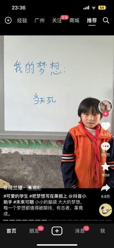 这是一张抖音平台的竖屏社交媒体截图，画面主体是一位穿着橙色校服、系着红领巾的小学生，他站在写有蓝色字迹的白板前，白板上写着“我的梦想：当天死”。下方的抖音配文却是充满正能量的话语：“#可爱的学生 #把梦想写在黑板上 @抖音小助手 #未来可期 小小的脑袋 大大的梦想，每一个梦想都值得被期待，有志者，事竟成。” 这里明显是小学生的笔误，把“当天使”写成了“当天死”，“使”和“死”发音相近，孩子书写时出现错误，形成了强烈的反差感，让人觉得既意外又好笑。