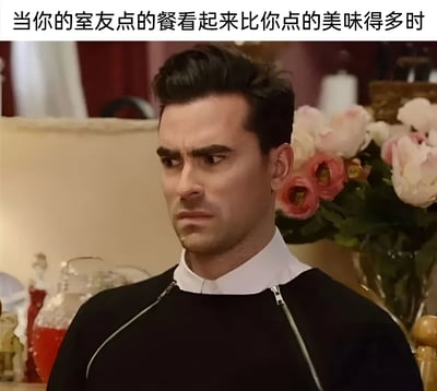 图片上方配有文字“当你的室友点的餐看起来比你点的美味得多时”，下方是一名男性角色的截图。他留着黑色短发，穿着黑色带拉链上衣，内搭白色高领衣物，表情严肃，眉头微皱，眼神侧视，流露出明显的羡慕或嫉妒情绪。背景中可见粉色和白色的鲜花以及一些室内陈设，整体营造出一种日常生活中因室友餐食更诱人而产生的微妙心理状态，场景轻松搞笑，容易引起共鸣。