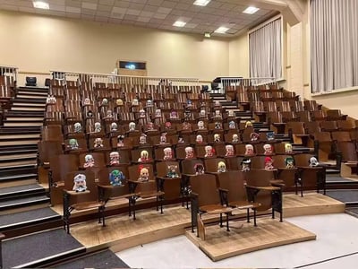 一张大学阶梯教室的照片，教室里排列整齐的棕色座椅上，几乎每个座位都摆放着一个小巧的毛绒玩偶。这些玩偶造型各异，颜色丰富，有不同的发型和服饰，仿佛一群迷你学生正在“上课”。教室环境宽敞，有多层台阶式座位、白色护栏、米色墙壁、灰色窗帘和方格天花板灯，整体氛围温馨又充满创意，像是一场可爱的玩偶主题活动或粉丝自发的趣味布置。
