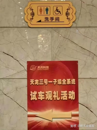 这是一张在大理石墙面上拍摄的现场指引标识照片，上方是带有男女及无障碍人士图标的洗手间指示牌，箭头指向左侧，标注有“洗手间”字样；下方是天兵科技的红色活动指示牌，印有品牌标识与英文“SPACE PIONEER”，核心文字为“天龙三号一子级全系统试车观礼活动”，箭头同样指向左侧，两个指示牌方向一致，呈现出活动现场的指引场景。