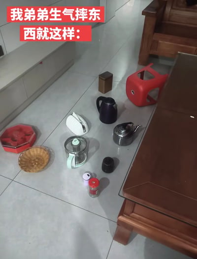 这是一张实拍的居家客厅场景图，浅灰色瓷砖地面上整齐排列着多种家用物品，包含红色八角果盘、竹编小筐、白色刀架、玻璃养生壶、金属茶罐、黑色烧水壶、银色小烧水壶、红色塑料凳，以及数个小型容器、调料罐等。图片顶部配有两行红底白字的标注文字：“我弟弟生气摔东西就这样：”，原本生气摔东西的预期和物品完好、整齐摆放的实际情况形成强烈反差，凸显出弟弟生气时也舍不得损坏家中物品的可爱感，颇具趣味性。