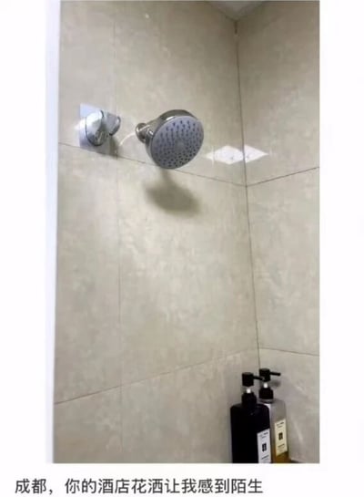 这张图片展示了成都某酒店卫生间内的场景，墙面铺着浅灰色的瓷砖，右下角台面上放置着两瓶洗护用品，最引人注意的是安装在墙面上的淋浴花洒——它并非紧贴墙面，而是通过一段管道向外凸出，与墙面形成了明显的空隙，这种违背常规的安装方式显得十分怪异。图片底部配有文字“成都，你的酒店花洒让我感到陌生”，直白地表达了拍摄者对这个奇葩酒店设施的吐槽与不解。