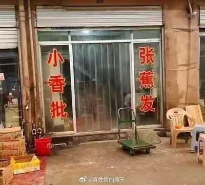 这是一张街边小店的实拍照片，店铺的玻璃推拉门两侧分别贴着红色的大字“小香批”和“张蕉发”，结合起来是谐音“小香蕉批发”，通过拆字重组的方式制造出类似人名的字样，形成搞笑的谐音梗效果。店铺外的地面上放着手推车、塑料椅，还有几个纸箱，看起来是一家做水果批发的街边小店。
谐音梗说明：将“小香蕉批发”拆分为“小香批”和“张蕉发”，把完整的经营内容拆成类似两个人名的组合，产生错位的搞笑感。