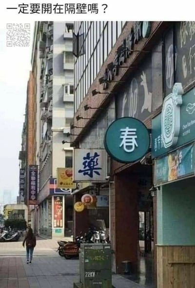 这是一张城市街头的实拍图，画面展示了街道旁的商铺区域，左侧是带有繁体‘藥’字的药店招牌，右侧紧邻的店铺招牌上有大大的‘春’字，两个招牌的文字组合在一起形成了谐音‘春藥’，图的顶部配有文字‘一定要開在隔壁嗎？’，用调侃的语气点出了这个巧合带来的趣味效果，让原本普通的店铺位置变得滑稽搞笑，这类偶然出现的文字组合常被作为趣味梗图在网络上传播。