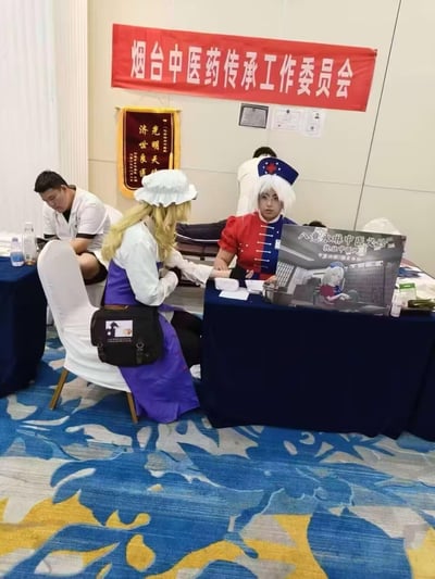 这是一场打破次元壁的趣味活动：在漫展现场，烟台中医药传承工作委员会开设了中医义诊摊位，工作人员cos成《东方Project》中的角色——八意永琳和雾雨魔理沙，以二次元的形式为漫展参与者提供中医诊脉服务。摊位背景挂着'烟台中医药传承工作委员会'的红色横幅与'光明天使 济世良医'的锦旗，旁边还有一位真实的医护人员在忙碌。这种将二次元cos与三次元中医便民服务结合的形式，既吸引了漫展观众的注意力，又推广了中医文化，充满趣味与新意。