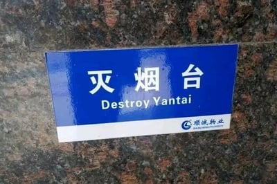 这是一块由顺诚物业设置的灭烟台标识牌，张贴在带有红黑相间纹理的墙面上。标识牌主体为蓝底白字，上方是中文“灭烟台”，指的是用于熄灭烟头的设施，但下方的英文却错误地译为“Destroy Yantai”，将“灭烟”的动作错误对应为“Destroy（毁灭）”，同时把“台”和城市“烟台”的拼音混淆，形成了一个极具搞笑效果的翻译错误，属于标识制作时的失误。