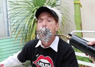 这是一张实拍的特技挑战照片，图中男子的嘴部被数十根烟卷塞满，烟嘴朝外呈密集放射状排列，他的一只眼睛紧闭，面部表情显得十分紧张。画面右侧有一只手握着喷火枪，枪口正对准这些烟卷，似乎即将点燃它们。男子穿着一件印有大嘴唇图案和英文“MOUTH”字样的黑色上衣，背景是户外的绿色植物与浅绿色房屋墙面，整体场景呈现出惊险刺激的特技挑战氛围，图中没有双关、谐音梗或暗示内容。
